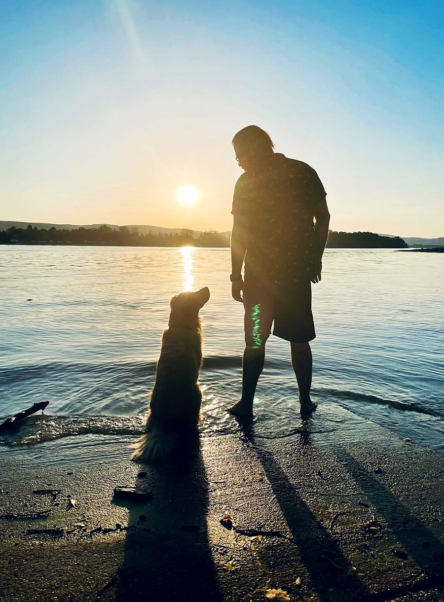Silhouette of Rick and his golden retriever Kenai standing at the edge of a river at sunset, with Kenai looking up at him.